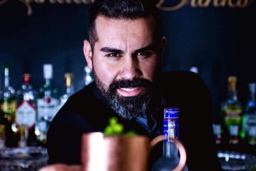 Homem sorridente misturando ingredientes em um copo para fazer um drink colorido em uma cozinha moderna.